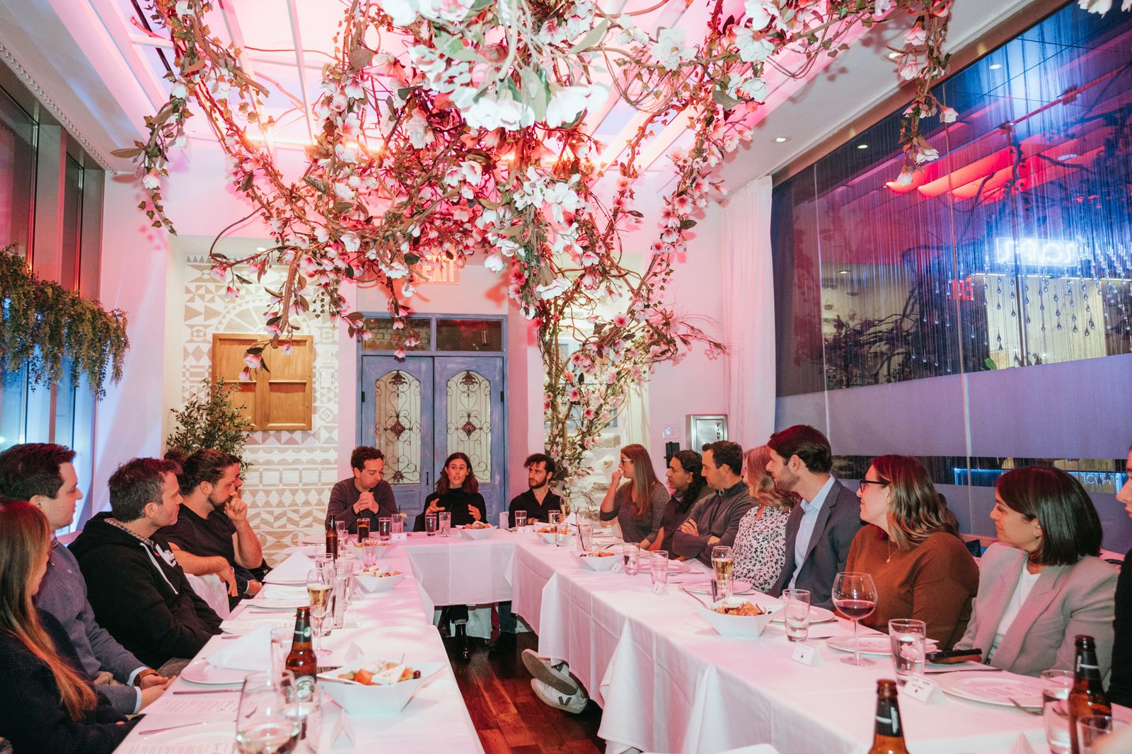 Guests seated around the table at a Dine & Deliver dinner, listening to someone speaking at the far end, under a canopy of blossom.