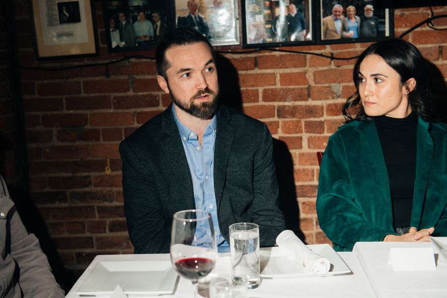 Two guests listening at the dinner table, against an exposed brick wall.