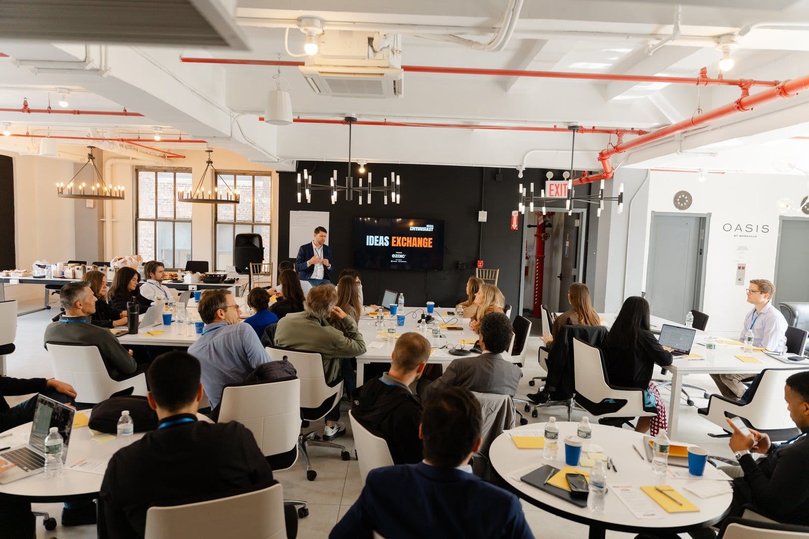 The room at work: thirty people at white tables under the chandelier, the Ideas Exchange screen at the far end.